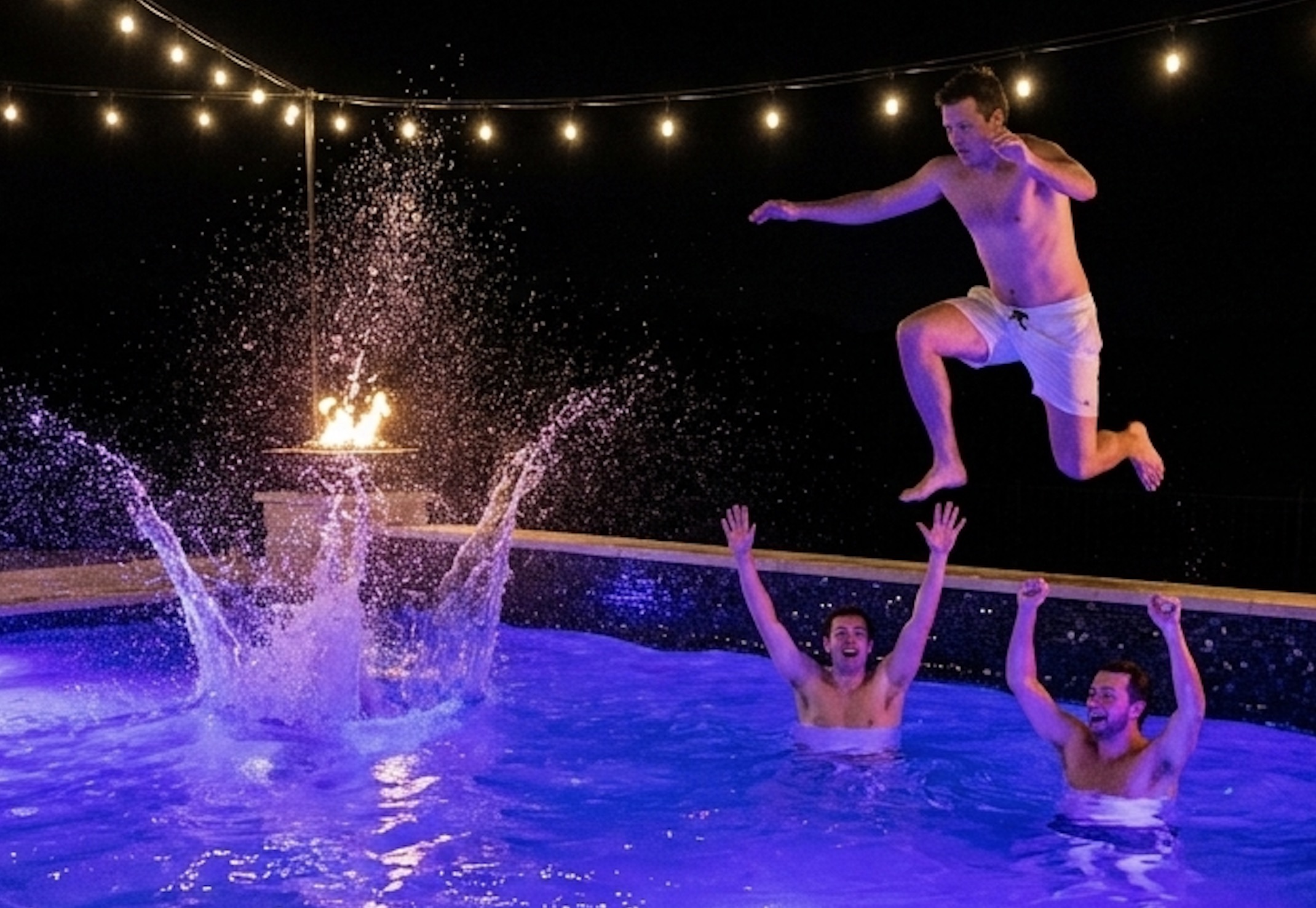 A pool lit at night beside a fire pit and string lights